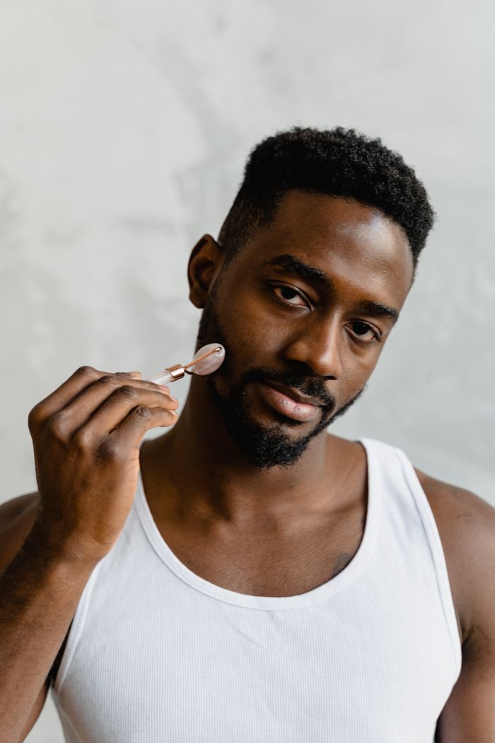 services-01 Portrait of a Black man using a jade roller for facial skincare in a white tank top.